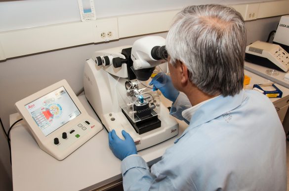 Grey-haired man in a white coat with blue latex gloves, sitting in front of a microtome (device for making very small incisions), photo from behind over his left shoulder.