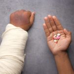 Zwei Arme, einer bandagiert, der andere hält bunte Pillen in der Hand.